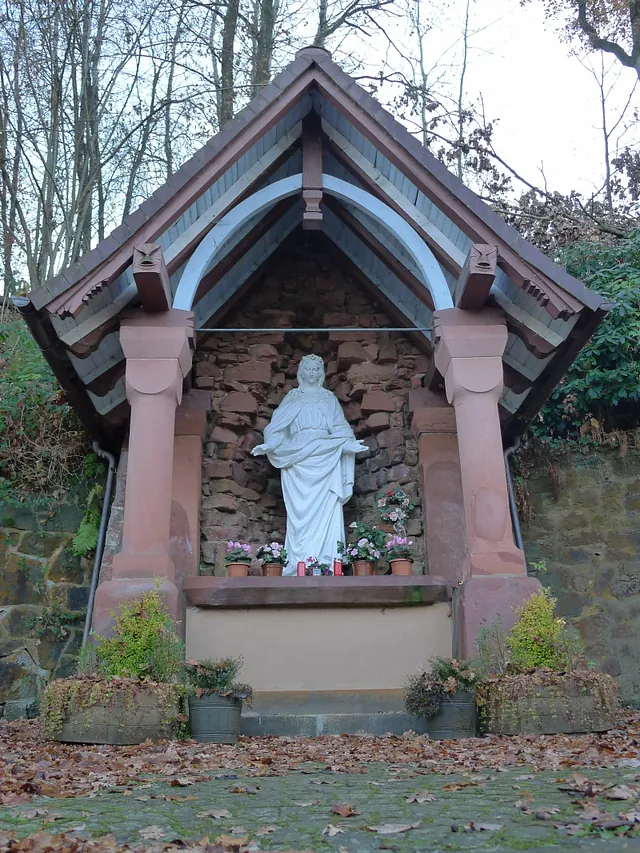 Weiße Marienstatue in einer überdachten Steingrotte mit Blumen.