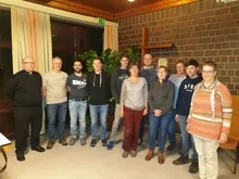 Pfarreigruppe mit Priester vor einem Holzkreuz stehend.